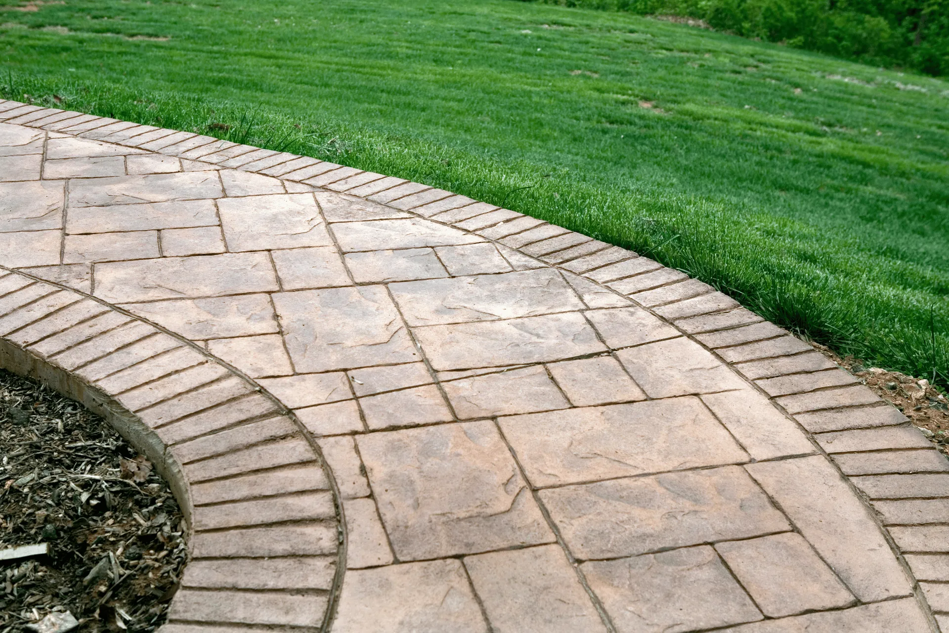 A curved pathway made of tan stamped concrete with a brick border, adjacent to a lush green lawn.