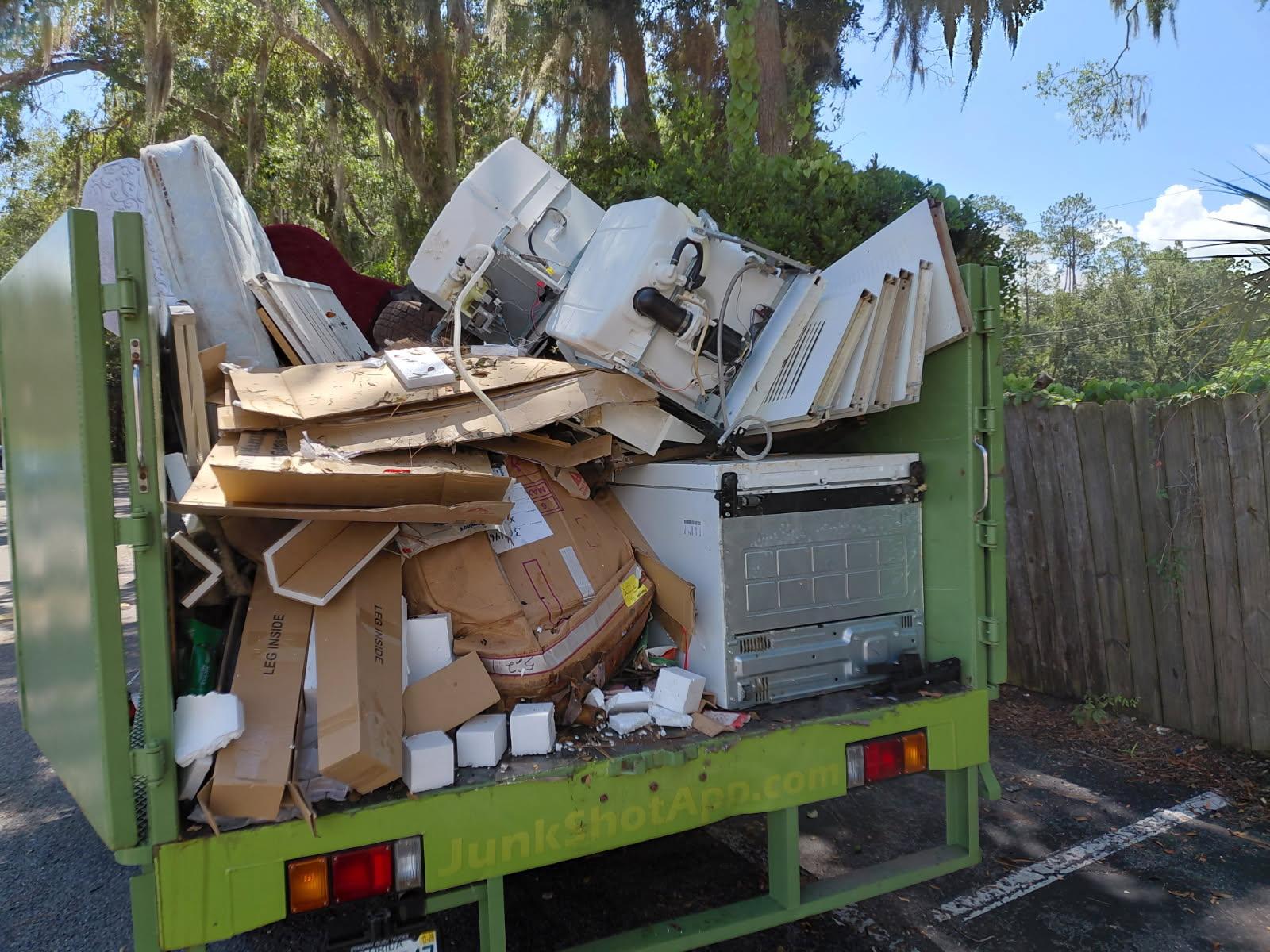 The back of a green JunkShot haul truck packed full with cardboard boxes, old appliances, and discar 