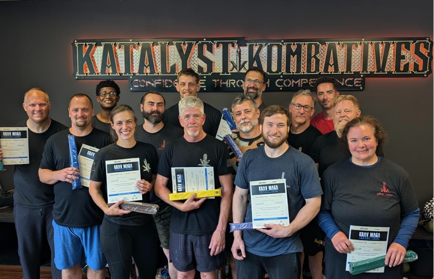 Adult Krav Maga students at Katalyst Kombatives in Virginia Beach gathered under the gym sign after a rank test, holding their diplomas and newly earned belts.