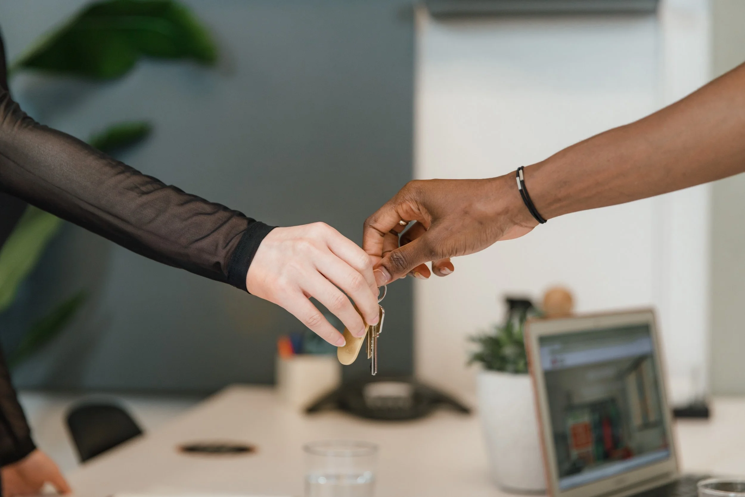 A close-up shot shows two hands exchanging a set of keys in an office environment. One hand, with fair skin and a sheer black sleeve, reaches out to receive the keys. The other hand, with dark skin and a black bracelet, holds the keys, with fingers delicately positioned around the keyring. The background is blurred, but suggests an office setting with a laptop, a plant in a white pot, and a grey wall with a hint of green foliage.