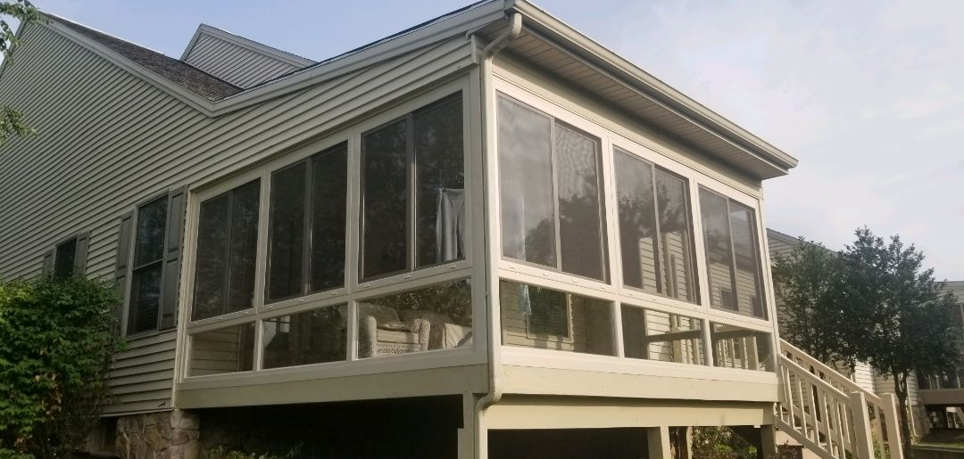 Elevated screened porch attached to a residential home, featuring large glass or mesh panels, white framing, and a sloped roof. The structure provides a bright, enclosed outdoor space for relaxation while protecting from insects and weather elements.