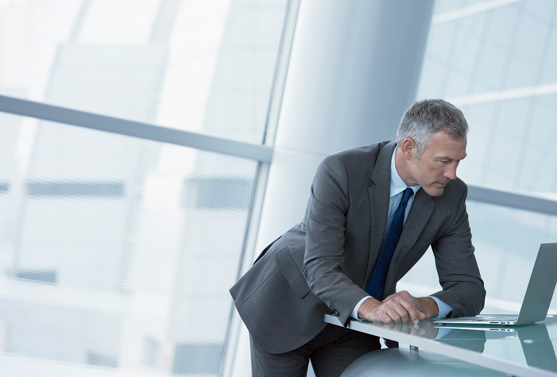 A man in a gray suit leans over a desk, focused on a laptop in a modern office with large windows. The scene conveys a professional and concentrated atmosphere.