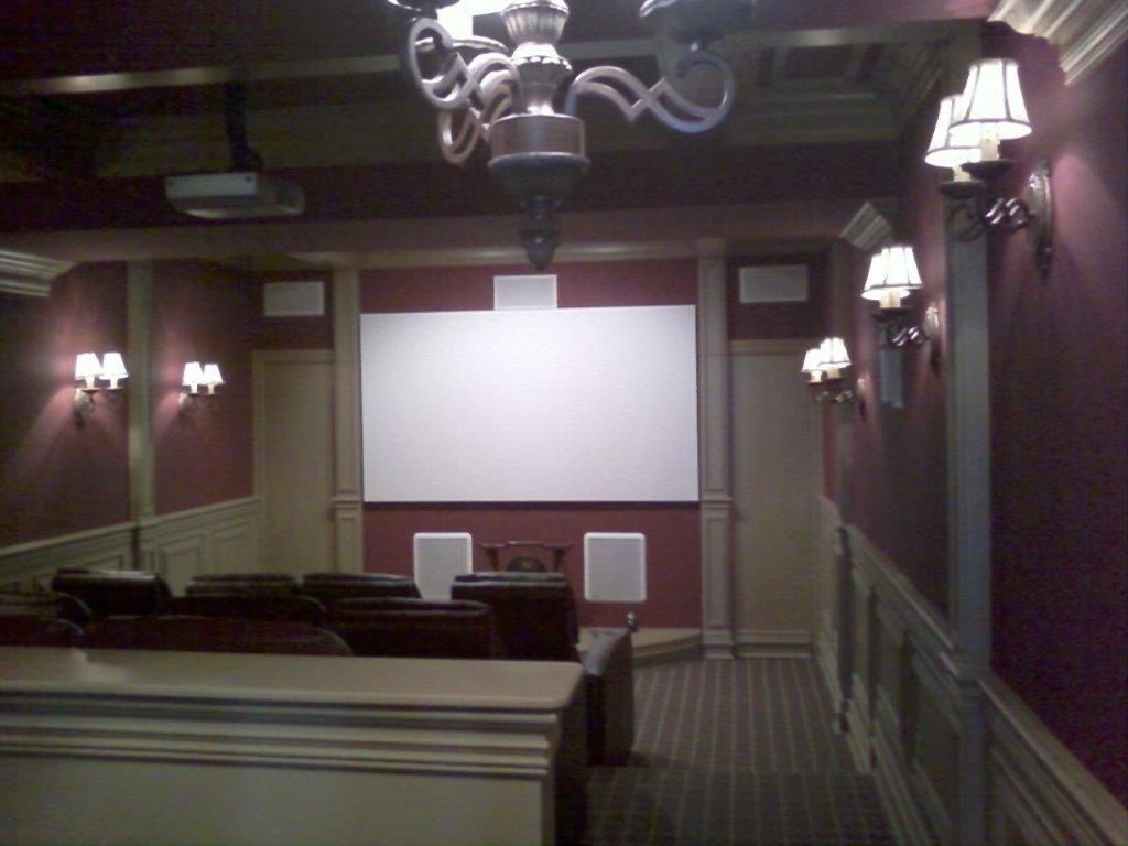 A cozy home theater room with a large projection screen, upholstered seating, wall-mounted sconces, and a chandelier. Rich red walls and decorative paneling create an elegant, cinematic viewing environment.