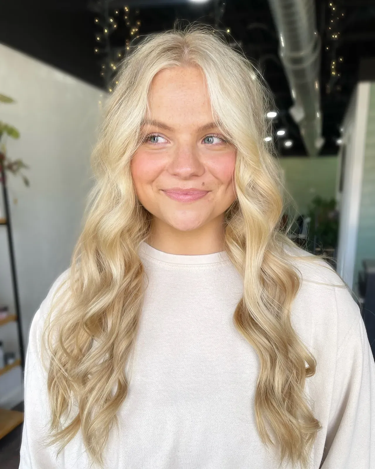 Woman with long wavy blonde hair and soft center part smiling gently, photographed inside a modern hair salon.
