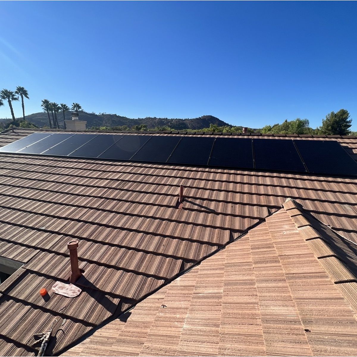 Clear view of a brown concrete tile roof equipped with a long row of sleek black solar panels under a bright blue sky, with surrounding trees and hills.