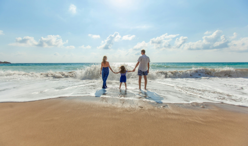 A family of three holds hands facing the ocean waves on a sunny beach. The sky is clear, evoking a sense of joy and tranquility.