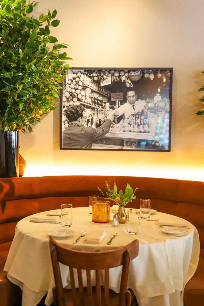 A cozy, curved velvet banquette table set for four, featuring a warm amber candle, fresh greenery, and a nostalgic black and white photo of an Italian market.