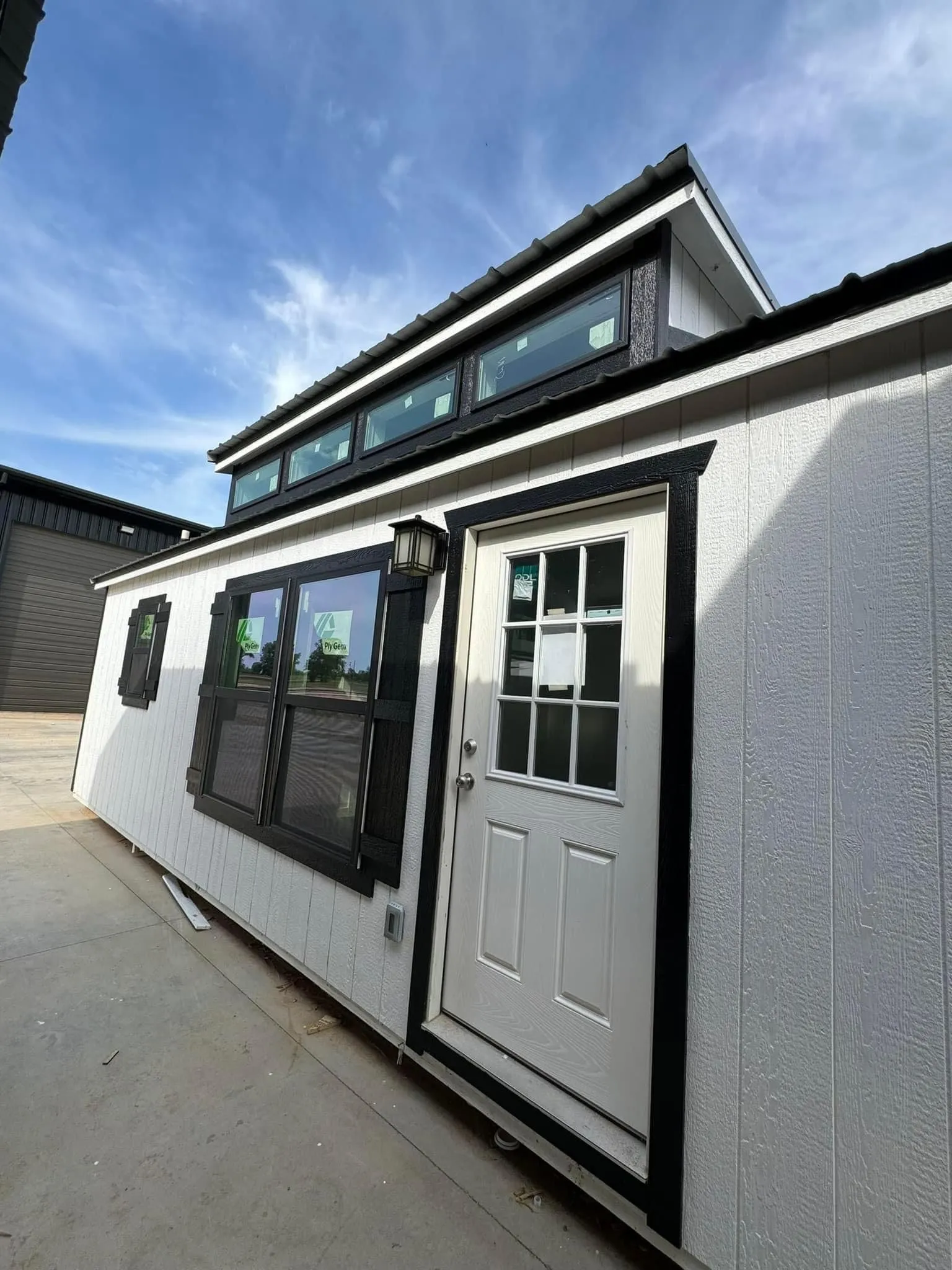 Modern white tiny home featuring black trim, clerestory windows, decorative shutters, and durable entry door. Compact design combines stylish appearance with practical living and efficient space utilization.