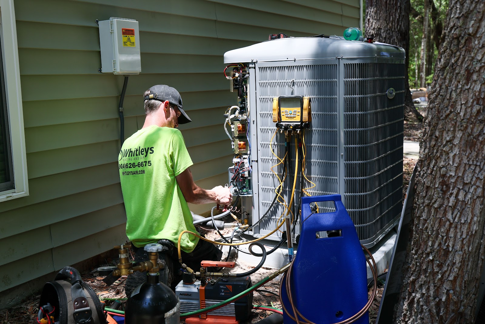 A technician wearing a bright green shirt with a company logo kneels to service an outdoor air condi 