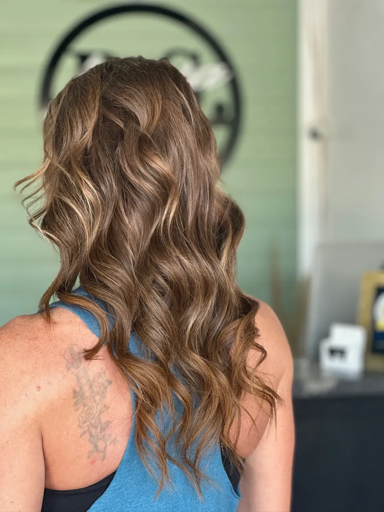 Woman with long soft wavy light brown hair styled with subtle highlights, photographed from back inside a modern salon setting.