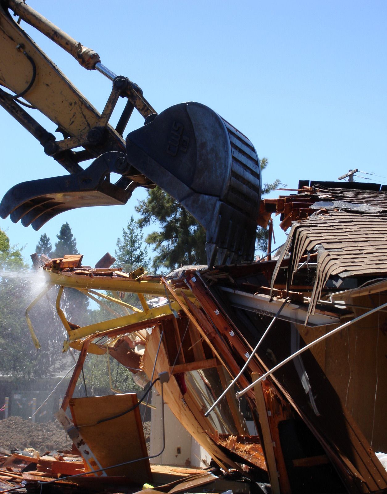 Close-up of excavator claw tearing through building materials, capturing active demolition, structural breakdown, and forceful removal of wood and roofing components during teardown process.