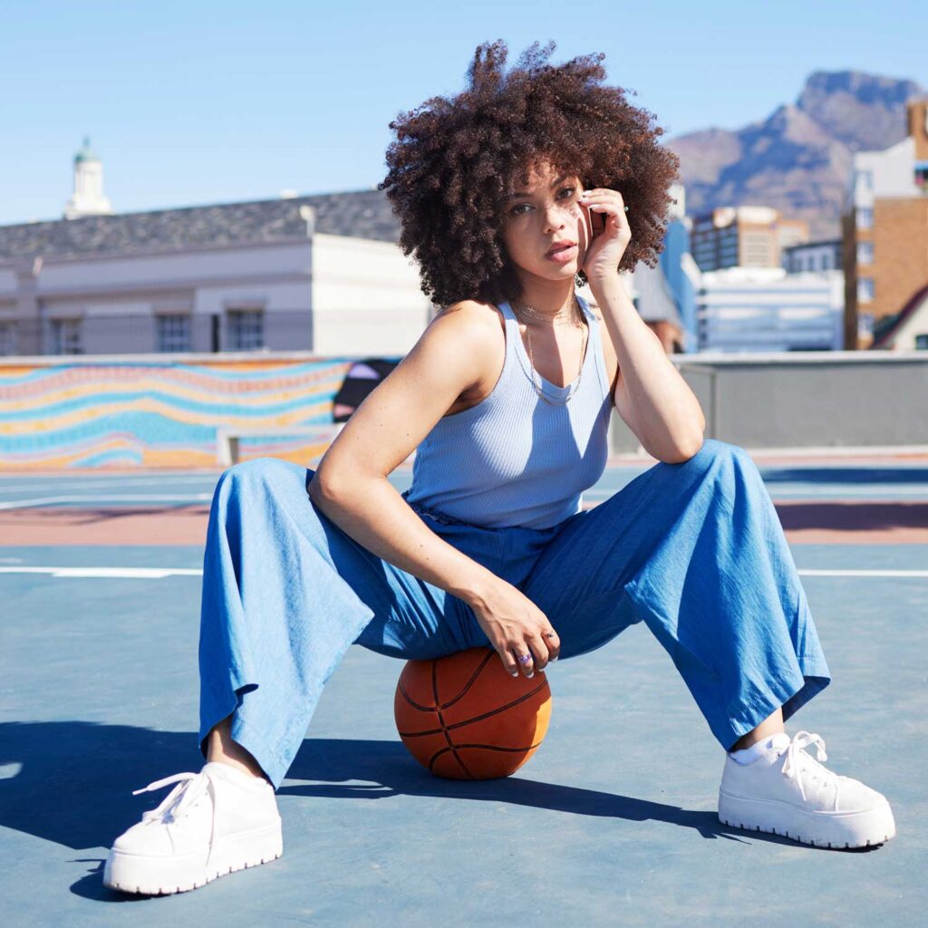 A young woman with voluminous curly hair sits on a basketball on a blue outdoor court. She is wearing a light blue ribbed tank top and loose-fitting blue pants. Her white sneakers have thick platform soles. In the background, there are buildings and mountains under a clear blue sky. A colorful mural decorates a wall behind her.