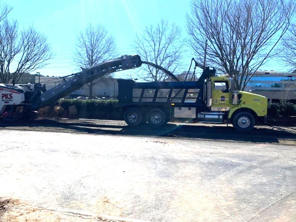 A road milling machine transfers milled asphalt through a conveyor into a yellow dump truck during pavement removal work. The construction equipment operates on a paved area with trees nearby.