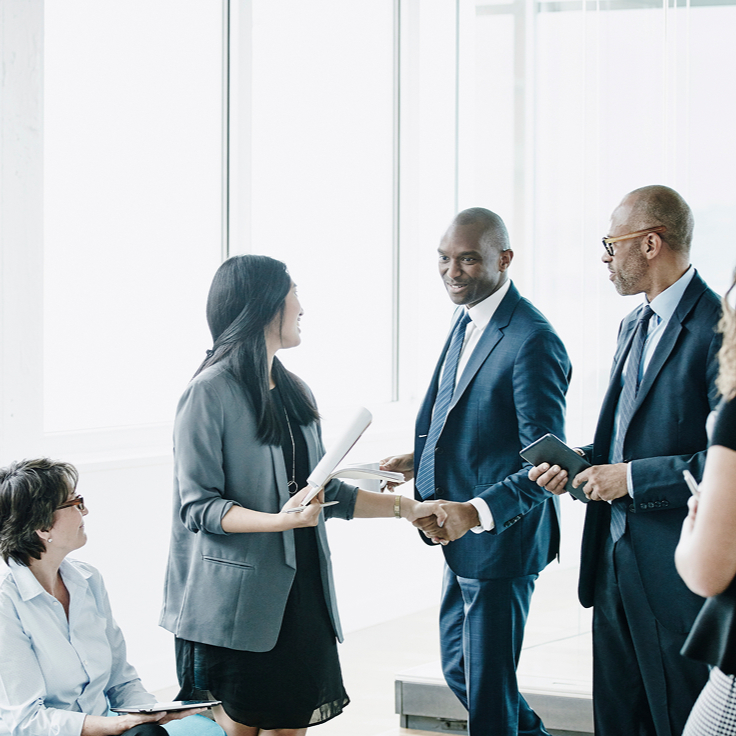 A group of diverse professionals in a bright office engage in conversation. Two men in suits shake hands, while a woman seated with a tablet looks on. The mood is collaborative.