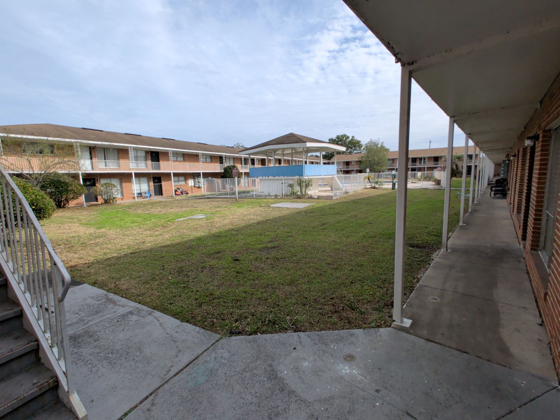 Apartment complex courtyard with grassy lawn, fenced swimming pool, and gazebo pavilion. Two-story brick residential buildings with exterior walkways and staircases surround shared outdoor community space.