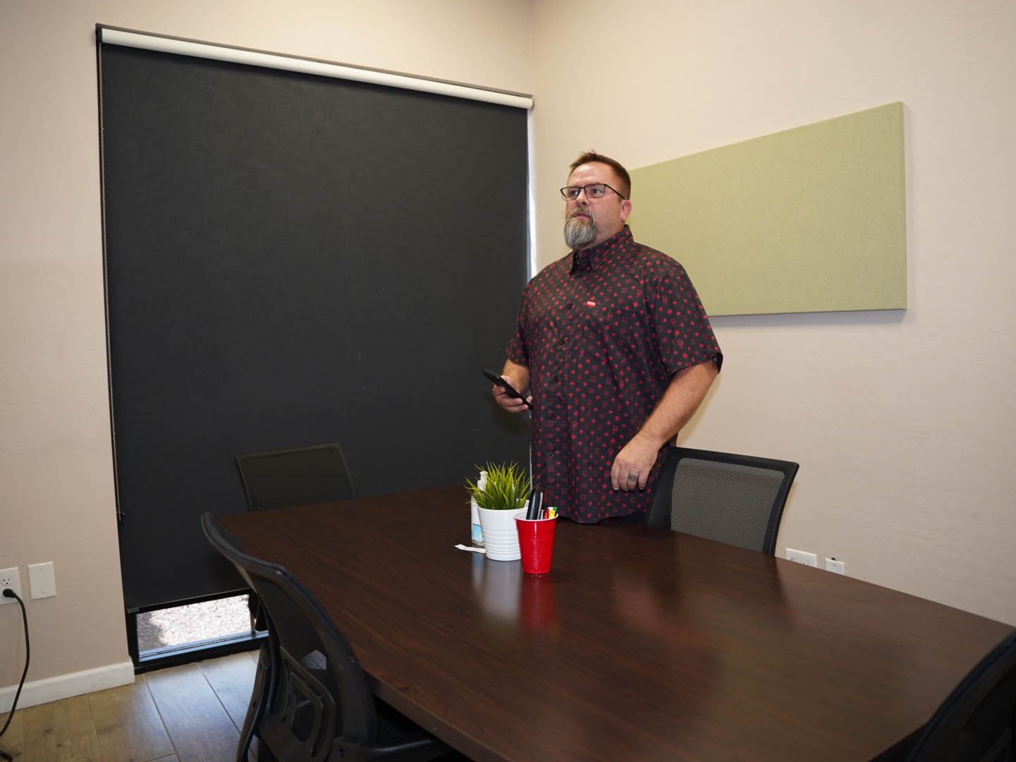 In this image, Mike Rux, a professional at Ciphers Digital Marketing, is seen standing at the head of a conference table during a session on social media marketing at their Gilbert, Arizona office. He is holding a phone and wearing glasses and a patterned short-sleeve shirt. The conference room features a black window shade, a potted plant, and office supplies arranged neatly on the table, creating a professional and organized environment. Mike's attentive and thoughtful demeanor highlights his engagement and dedication to the discussion.