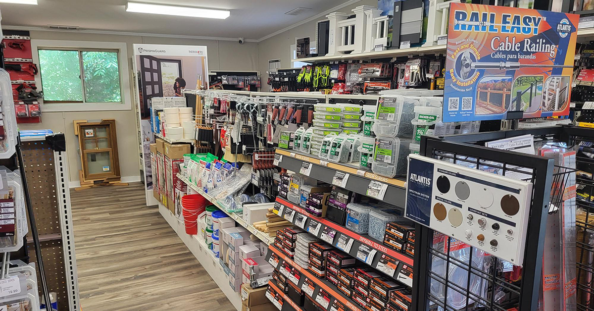 Interior view of a Builders FirstSource showroom aisle stocked with hardware and building supplies, including deck screws, sandpaper, paint supplies, and drywall fasteners on labeled shelving. A Rail Easy cable railing display and an Atlantis Rail color sample board are mounted at the end of the aisle, with sample windows and millwork displayed against the back wall under fluorescent lighting.
