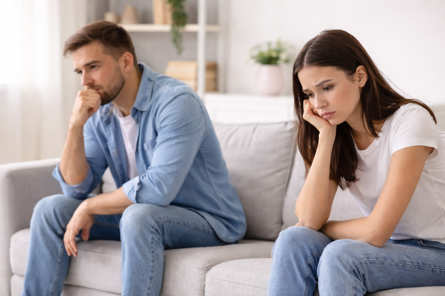A man and a woman sit on a couch with their backs to each other, looking dejected and deep in thought. The man wears a blue denim shirt and jeans, while the woman wears a white t-shirt and jeans. The background shows a shelf with books and a potted plant, suggesting a living room setting. The overall mood is somber and reflective, possibly indicating relationship troubles.