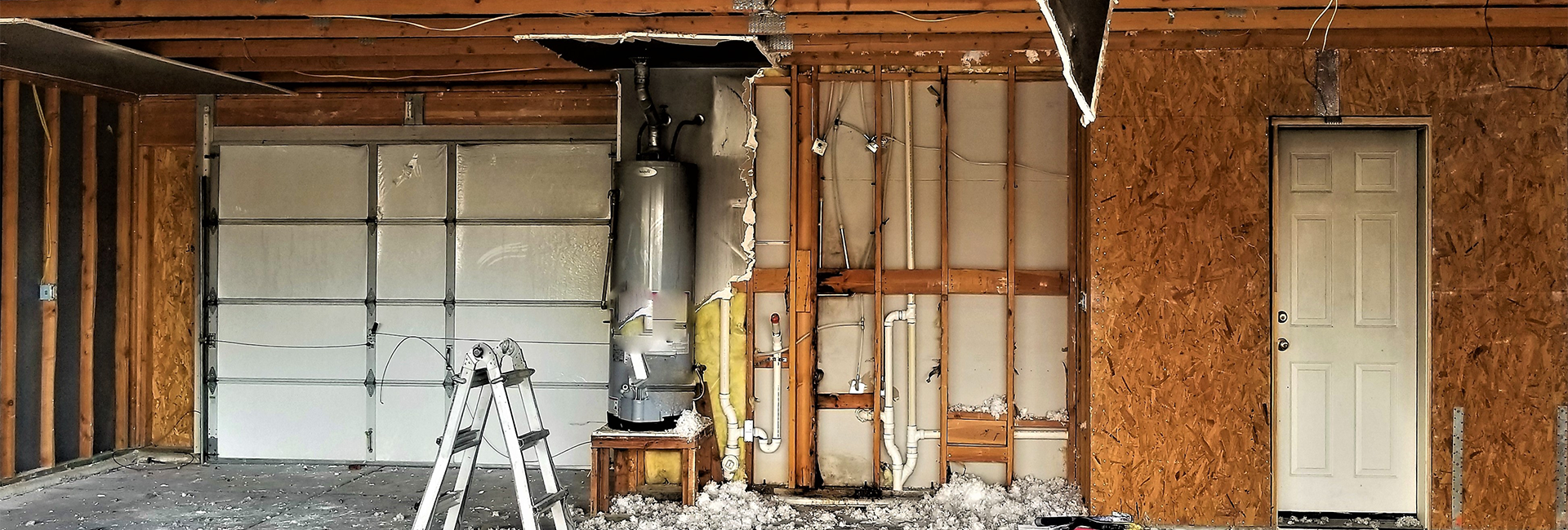A partially demolished garage interior features a white garage door on the left and a white interior door on the right, both set within exposed wooden studs and plywood walls. A large silver water heater stands to the left of the interior door, next to a silver stepladder. Debris, including insulation and wood scraps, is scattered across the concrete floor. Electrical wires and plumbing pipes are visible along the unfinished walls.