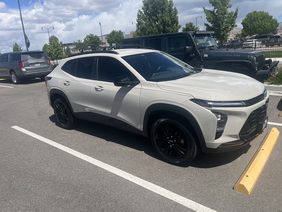 A light-colored Chevrolet Trax is parked in a parking lot next to other vehicles. The car has black rims and tinted windows. A yellow parking bumper is in front of the car. In the background, there is a black Jeep Wrangler and a grey SUV, along with some trees and buildings under a partly cloudy sky.