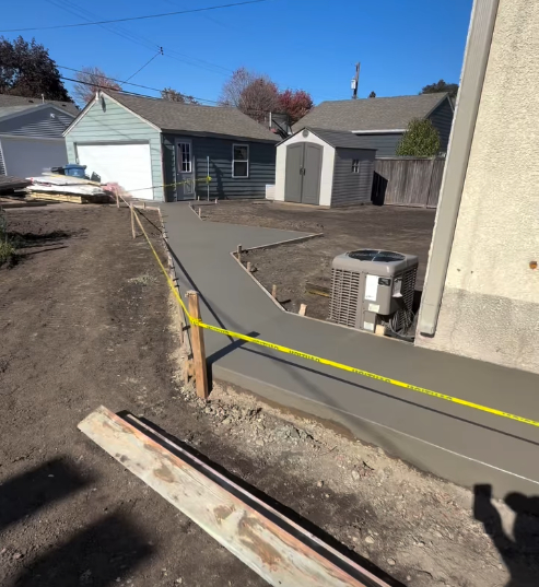 n angled outdoor view showcasing a freshly poured, smooth gray concrete walkway project under construction. The path winds along the side of a home past an air conditioning unit toward a detached garage and storage shed in the backyard. Wooden framing stakes and bright yellow safety caution tape line the perimeter of the drying concrete.