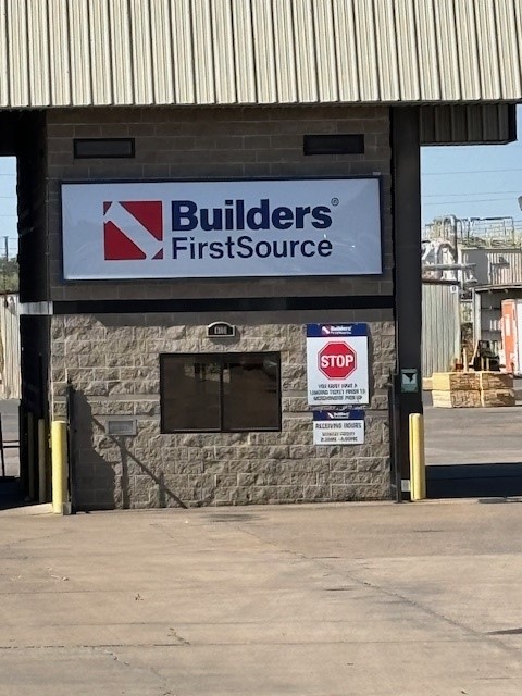 A close-up view of a Builders FirstSource entrance gate and receiving area. The sign with the Builders FirstSource logo is prominently displayed above a small window in a stone-clad structure. Below the sign, there are additional instructions, including a red stop sign that reads "STOP: Your first stop is receiving office." The receiving office hours and instructions are also posted on a sign. In the background, part of the lumber yard is visible, with stacks of wood and industrial equipment. The area is well-lit with a clear, sunny sky.