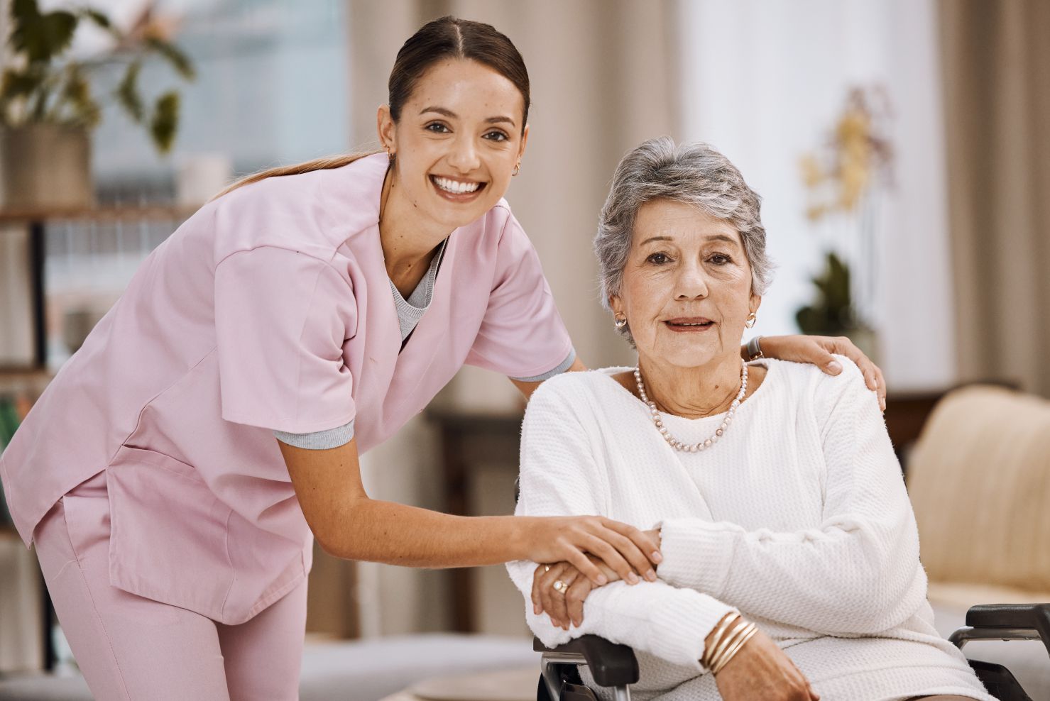 A smiling young caregiver in a pink uniform places her hands on the shoulders of an elderly woman sitting in a wheelchair. The caregiver is leaning forward and smiling directly at the camera. The elderly woman, wearing a white sweater and pearl necklace, looks forward with a gentle expression. The background is blurred, suggesting an indoor setting like a home or care facility, with hints of furniture and plants.