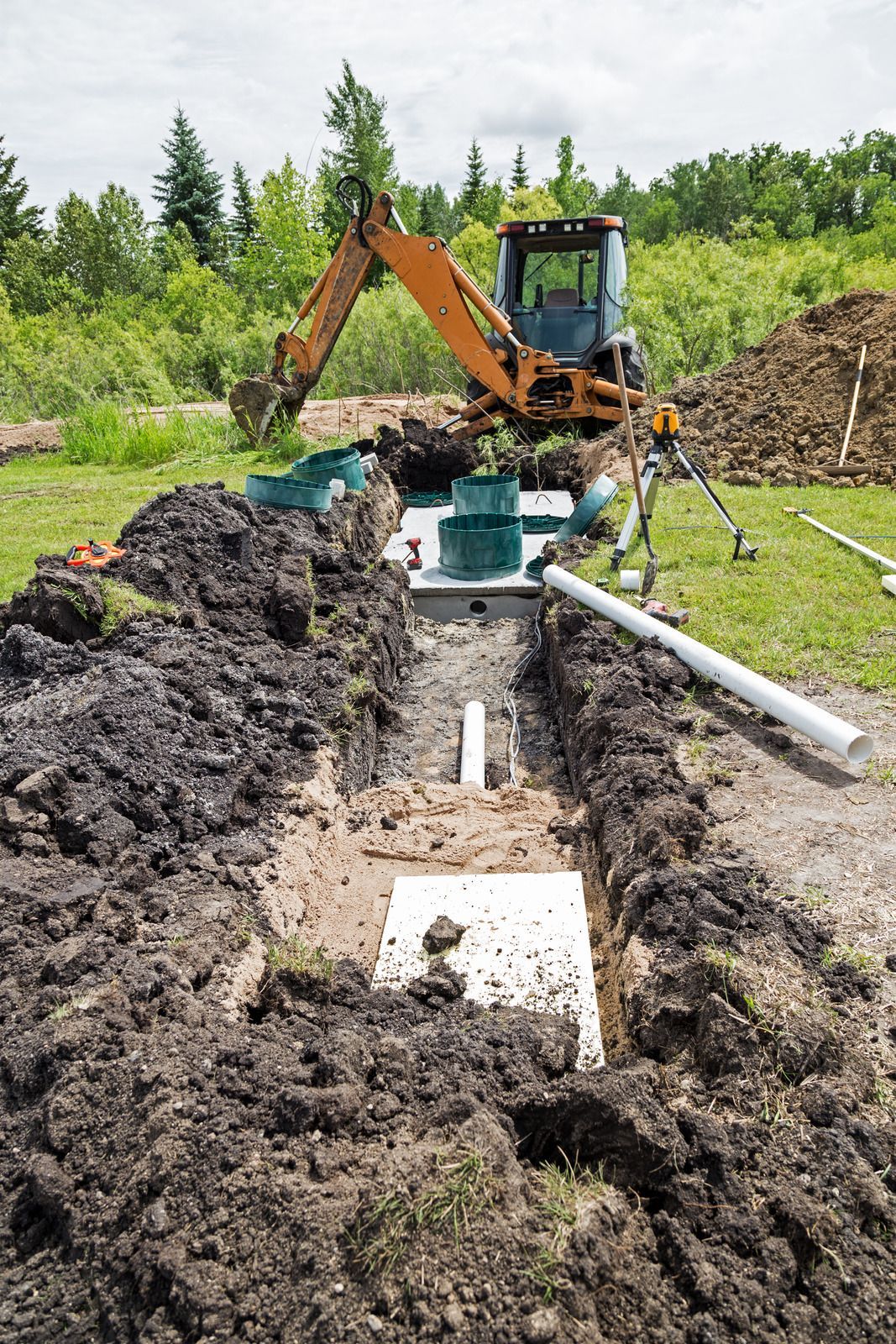 An orange backhoe works at a residential installation site where a septic system is being placed in a long dirt trench. Green risers, white PVC piping, and leveling tools are visible.