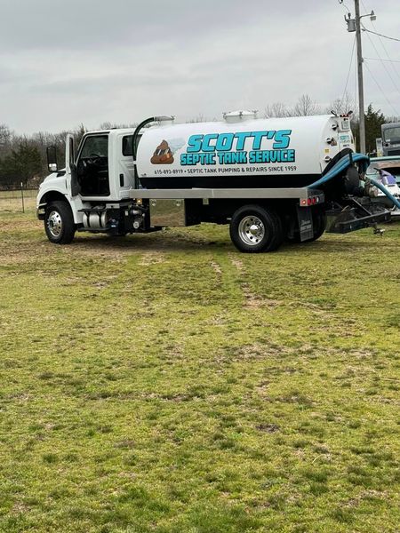 A professional white vacuum truck for Scott&rsquo;s Septic Tank Service parked in a grassy field. The truck features a large tank with blue branding, a phone number, and a humorous mascot.