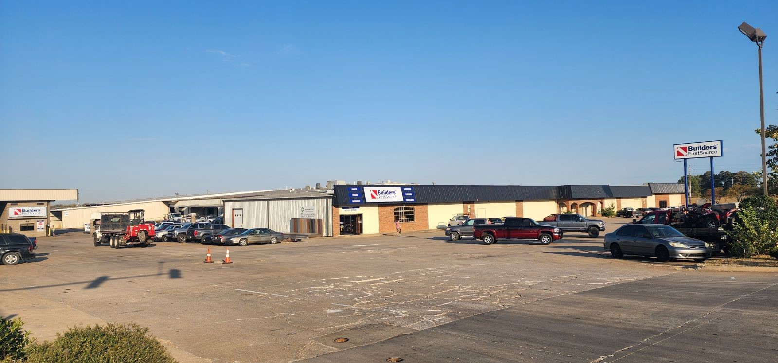 A wide-angle view of a Builders FirstSource facility, including a parking lot and storefront. The building features a mix of brick and metal siding with large signage displaying the Builders FirstSource logo above the entrance and on a standalone sign to the right. Several vehicles, including trucks and a red dump truck, are parked in the lot. The facility appears to include multiple sections, with a lumber yard visible in the background. The sky is clear and blue, and the overall scene reflects a busy, industrial retail location.