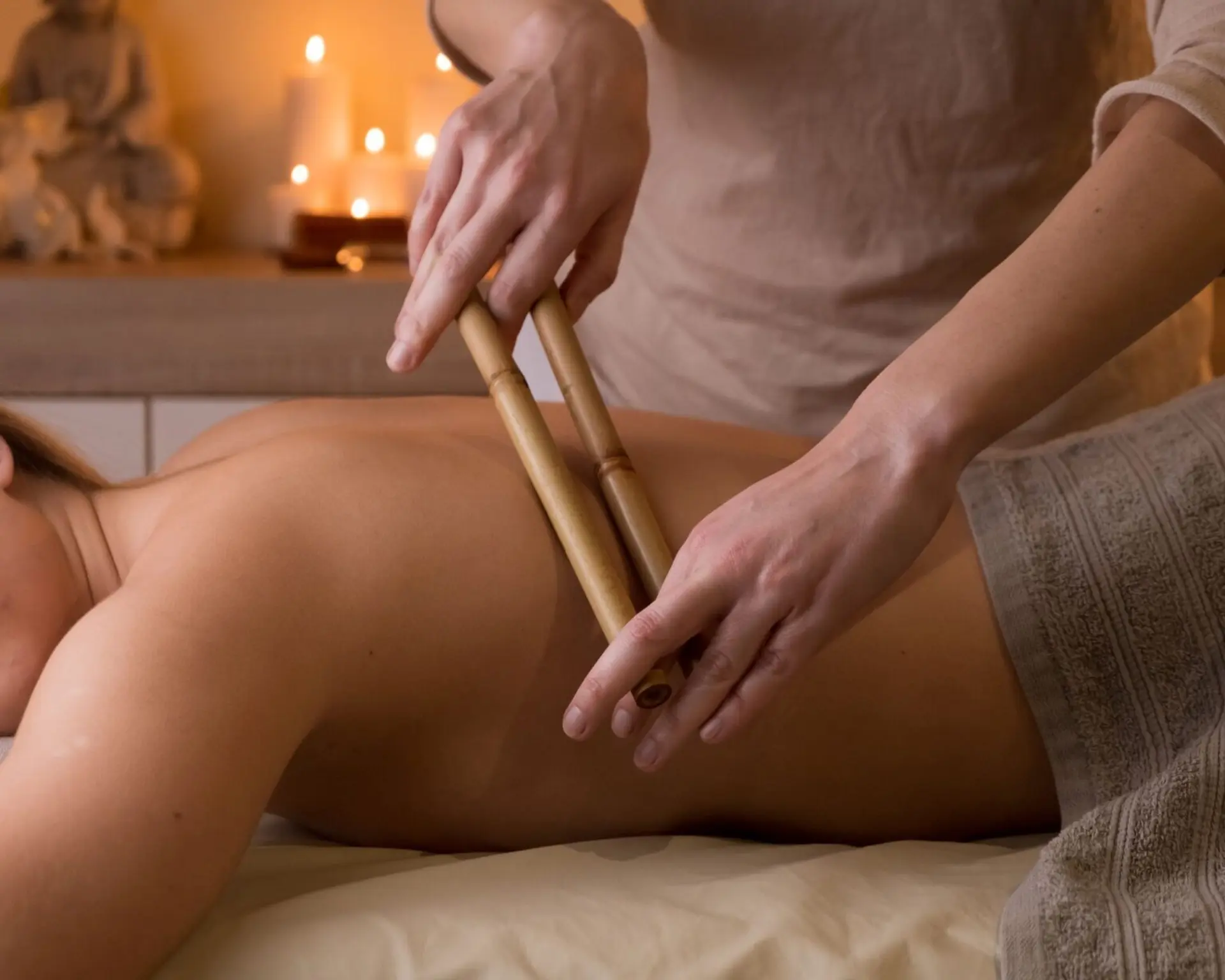 A close-up shot shows a massage therapist using bamboo sticks to massage a person's back. The scene is dimly lit and features candles and a statue in the background, suggesting a spa or wellness setting.