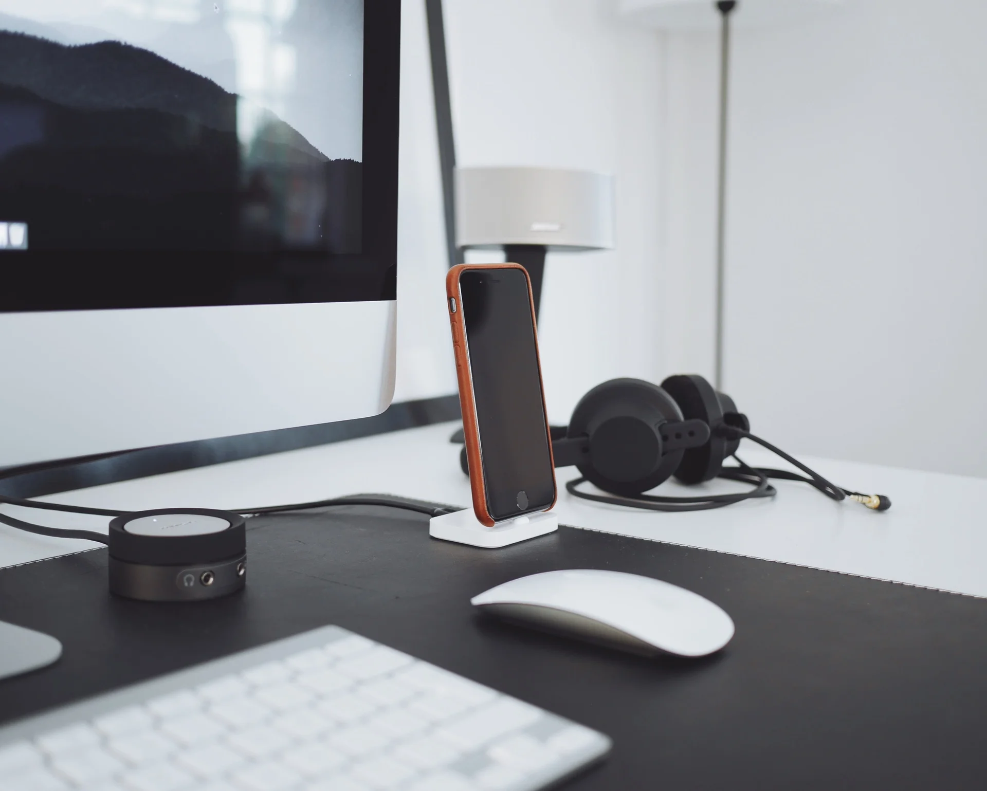 A modern home office setup featuring a computer monitor displaying a mountain landscape, a smartphone on a charging stand, a pair of black headphones, a white computer mouse, and a white keyboard, all arranged on a black desk mat with a white desk surface.