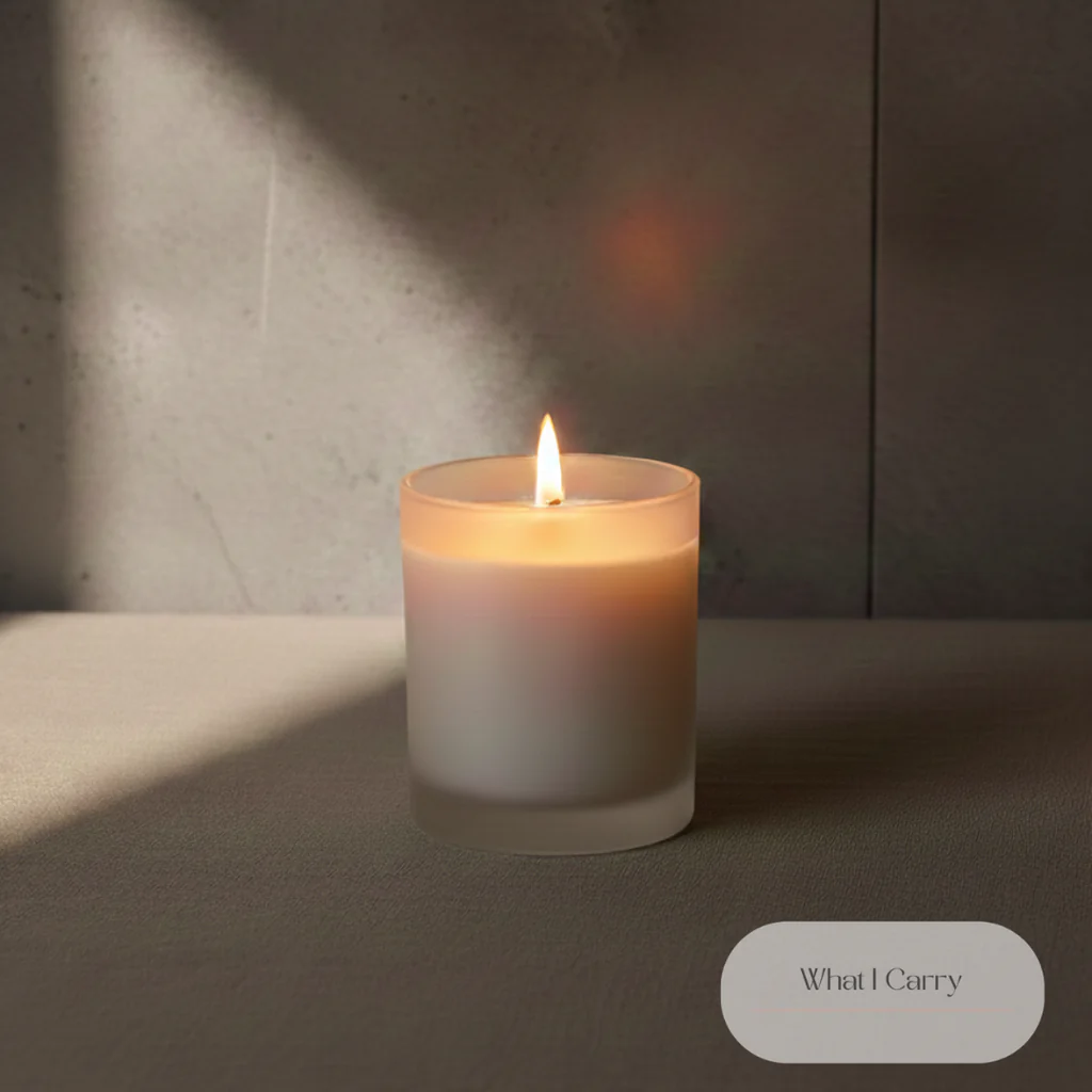 A lit candle in a frosted glass container sits on a textured surface, with soft light and shadows casting on a gray tiled wall in the background. The candle flame is bright and warm, illuminating the wax within the container. In the bottom right corner, there is an oval-shaped graphic with the text "What I Carry" written in elegant script.
