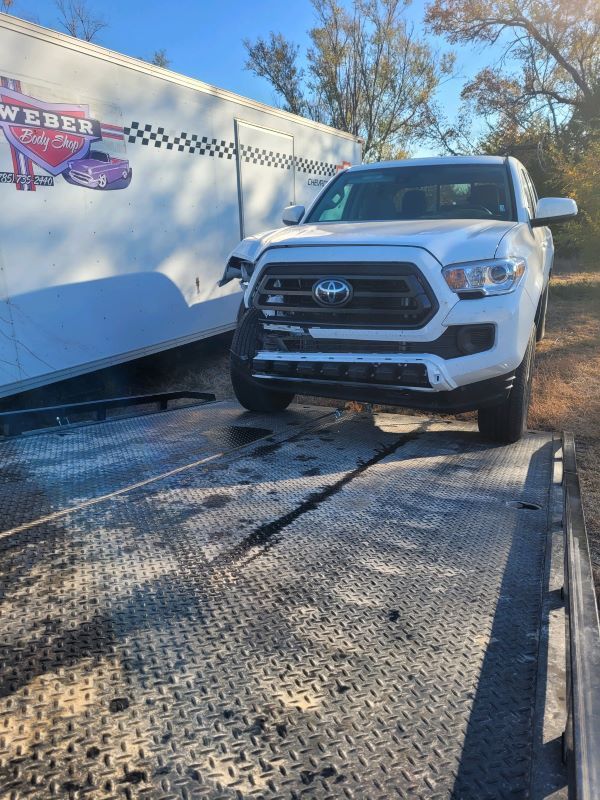 A white Toyota Tacoma pickup truck with heavy front-end passenger side damage is being pulled by a steel winch cable onto the metal diamond-plate bed of a flatbed tow truck.