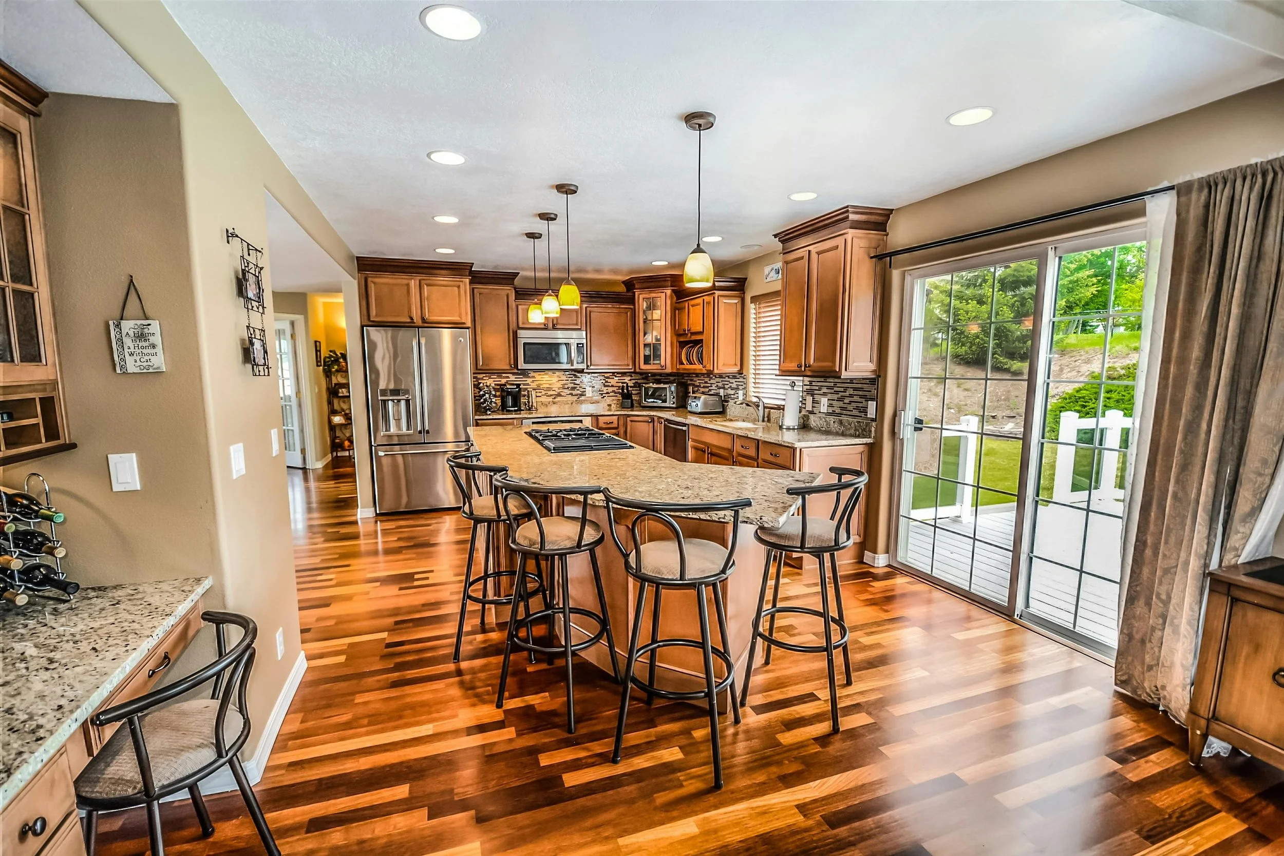 A spacious, modern kitchen featuring a large island with bar stools, stainless steel appliances, and a sliding glass door that opens to a backyard patio.