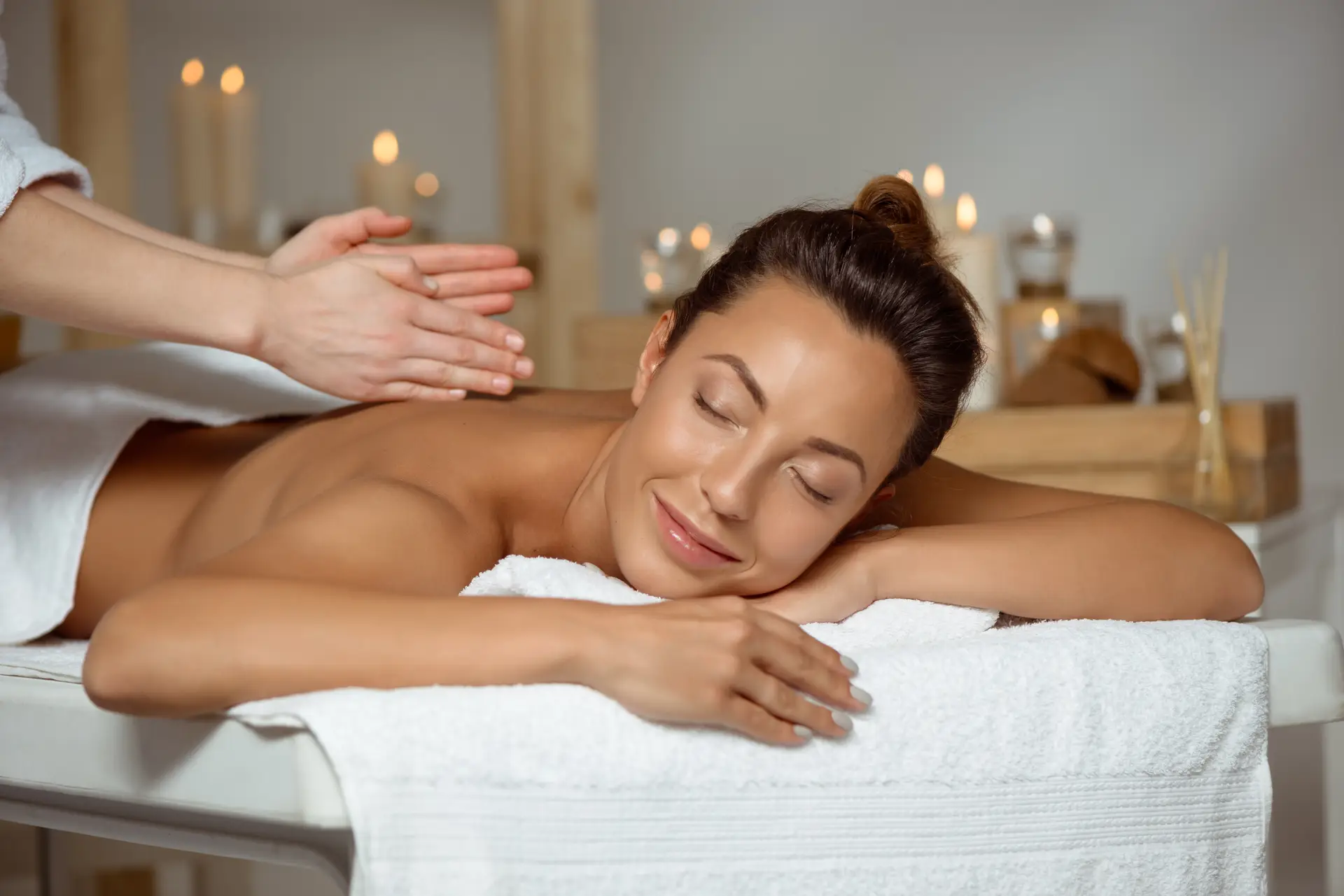 A woman lies face down on a massage table, receiving a back massage. Her eyes are closed and she has a relaxed expression. The room is softly lit by candles, and there are spa-like decorations in the background, including wooden shelves with bottles and reeds.