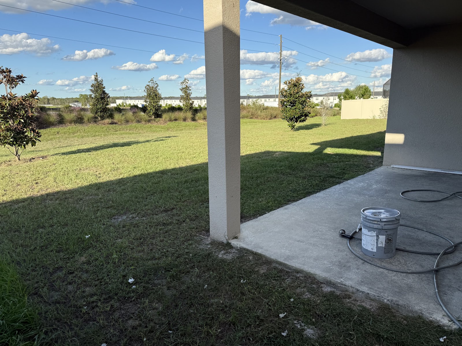 A view of a backyard with a concrete patio, a grassy lawn, and several trees under a blue sky with white clouds. A bucket with a spray nozzle and hose is on the patio.