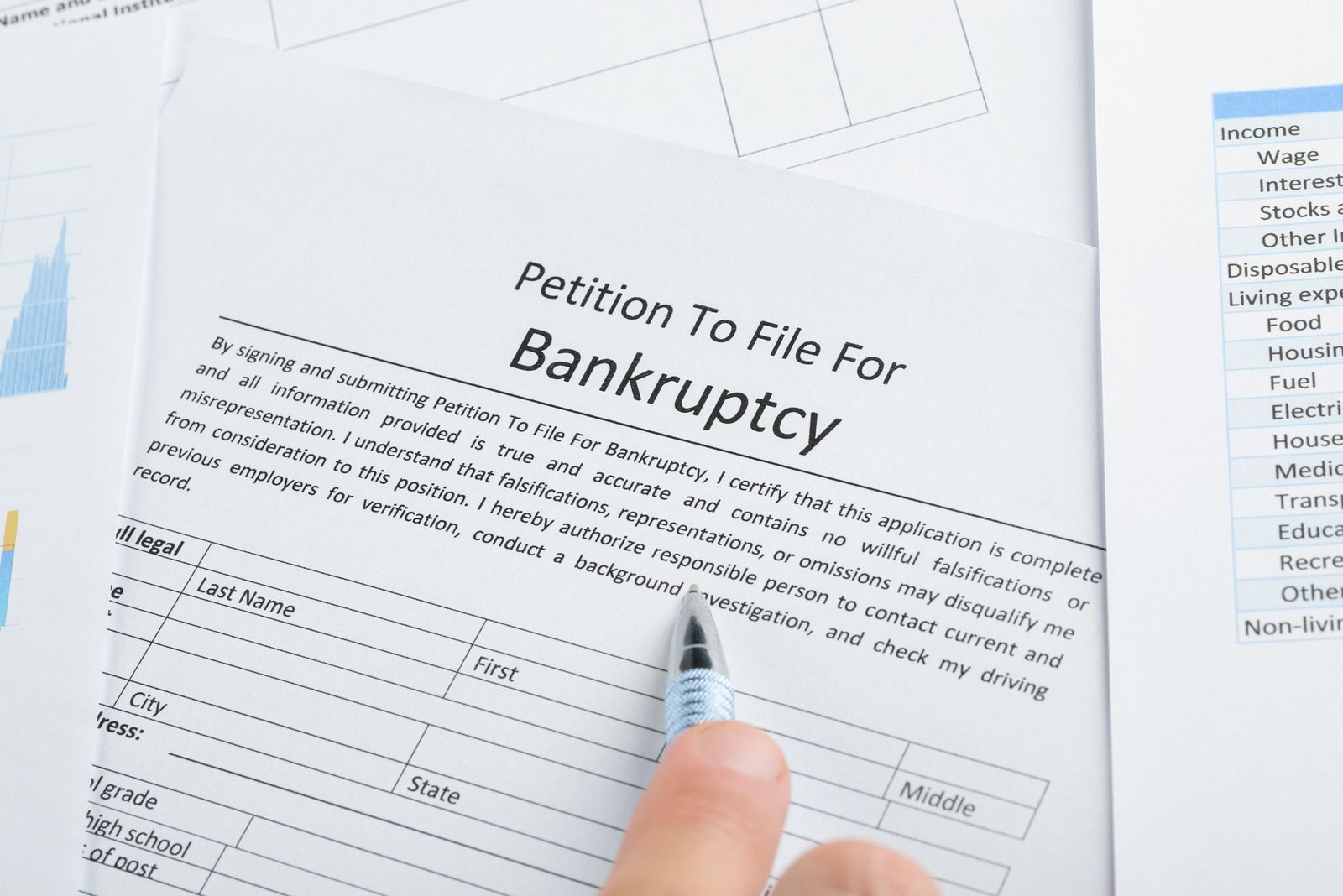 A hand holds a silver pen pointing at a "Petition To File For Bankruptcy" form with name and address fields, surrounded by financial charts and an income and expense worksheet.