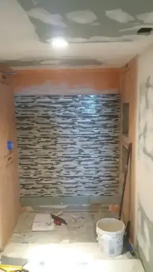 A bathroom under construction shows a shower area with horizontally striped gray and white tiles. The walls are unfinished, and a white bucket is visible.
