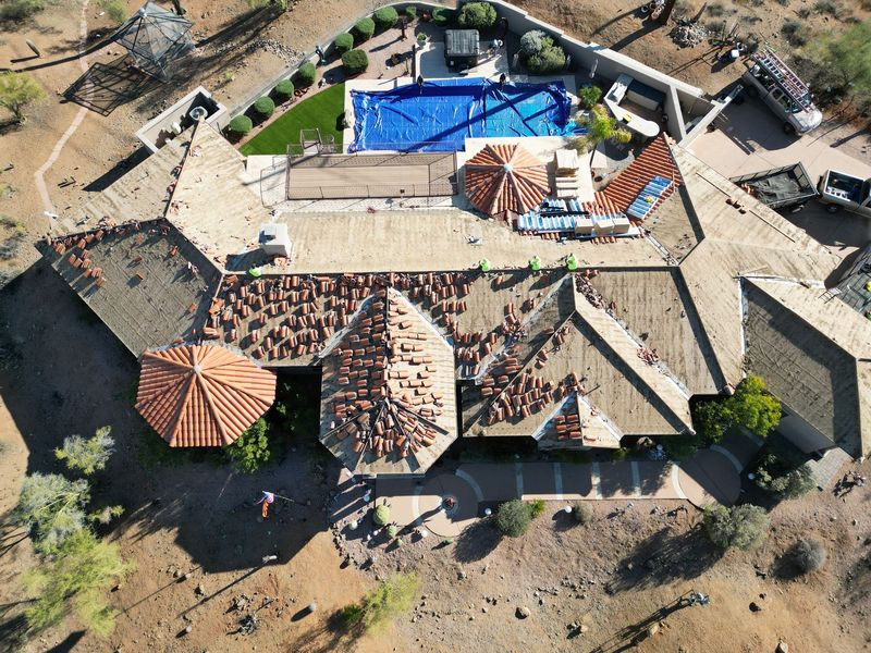 A bird's-eye aerial view captures a large desert home undergoing a roof replacement. Workers are actively laying red clay tiles across expansive roof sections next to a blue-covered backyard pool.
