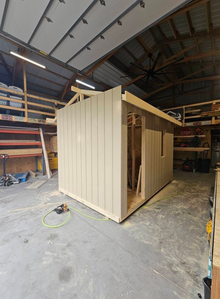 A light tan-sided storage shed is under construction inside a large workshop or garage. The walls and floor are mostly complete, with the exposed roof rafters and unfinished framing visible, along with tools nearby.