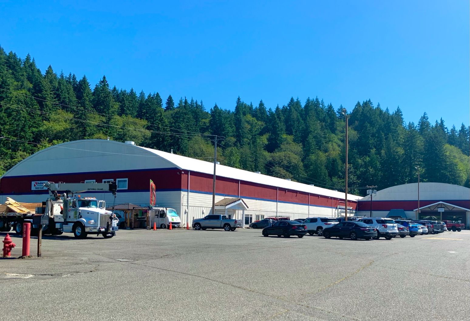 A wide view of the Builders FirstSource facility in Issaquah, WA, featuring a red and white building surrounded by a parking lot with various cars and trucks. The facility is backed by a lush green forest under a clear blue sky.