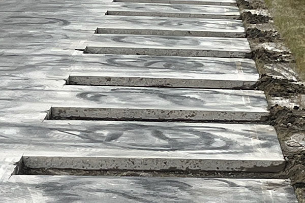 A wide-angle, low-perspective photograph taken along the longitudinal joint of a concrete highway. A series of parallel, rectangular slots have been precisely saw-cut and excavated into the existing concrete pavement perpendicular to the joint. These clean slots are empty and ready to receive dowel bars as part of a Dowel Bar Retrofit (DBR) load transfer restoration process. The adjacent pavement shows grinding residue and tire marks.