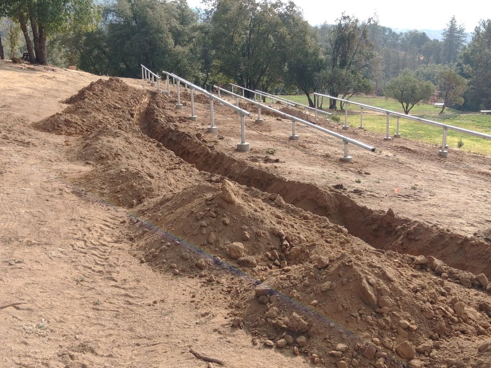 Long excavated trench runs across a sloped construction site beside metal rail structures set on concrete footings, with loose soil piles, trees, grassy areas, and wooded hills surrounding the project.
