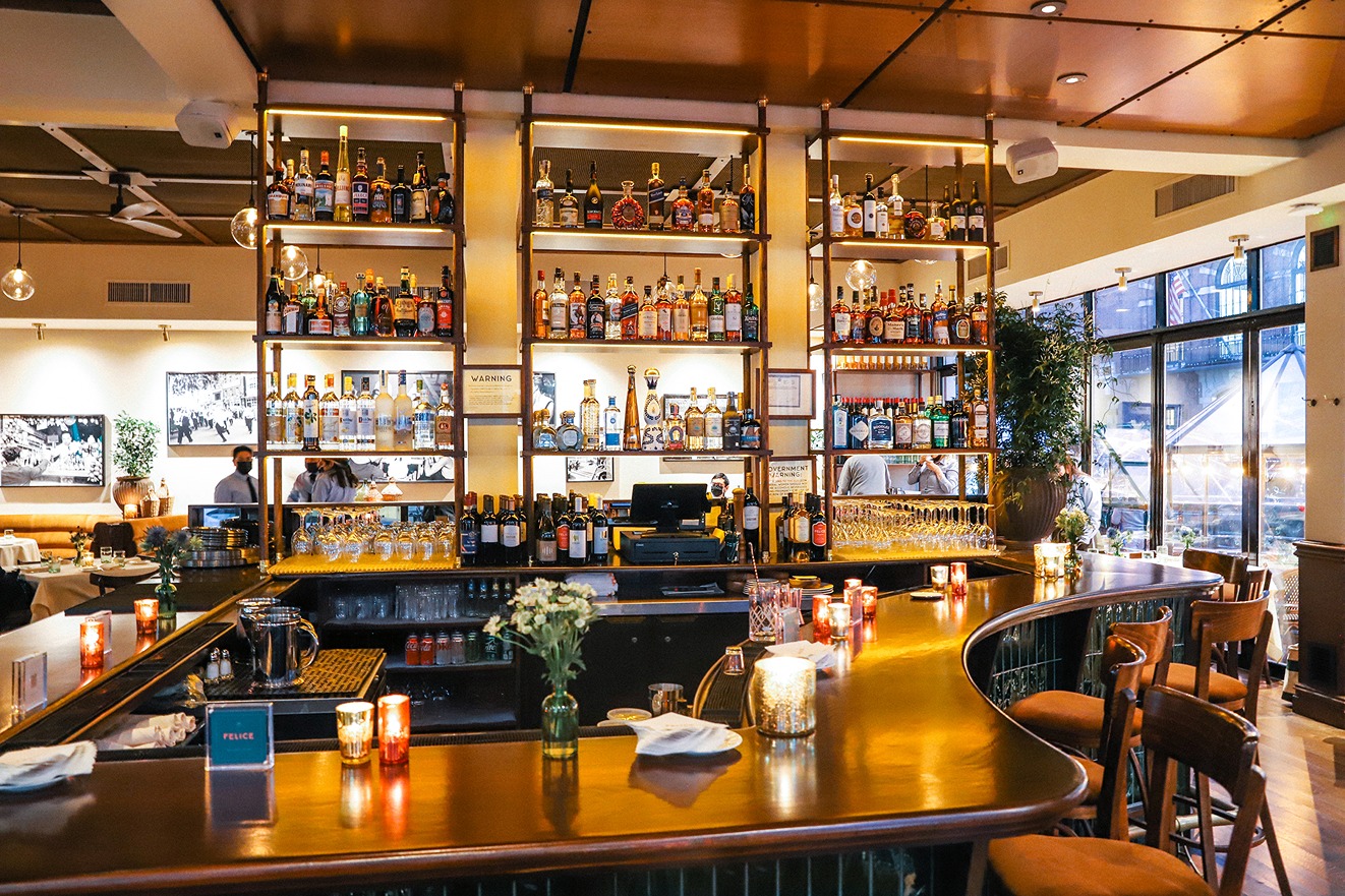 A warm, sophisticated bar with a curved wood countertop, glowing candles, and extensive liquor displays under soft ambient lighting.