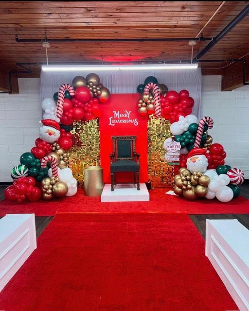 Festive Christmas photo backdrop featuring red carpet, Santa chair, balloon garlands in red, green, white, and gold, candy cane accents, glitter panels, holiday signage, creating a cheerful seasonal event setup.