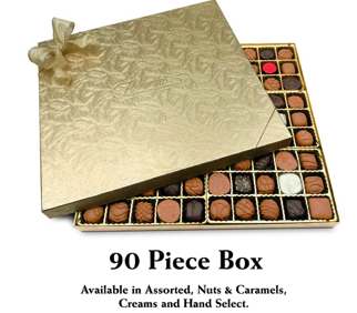 This photograph features a 90-piece box of Florence's Exquisite Chocolates, likely from their Rexburg, Idaho location. The chocolates are packaged in a luxurious gold box, tied with a large, elegant bow. The lid of the box is partially open, revealing a meticulously arranged assortment of chocolates. The chocolates vary in type, including nut clusters, caramels, creams, and other specialty pieces.
