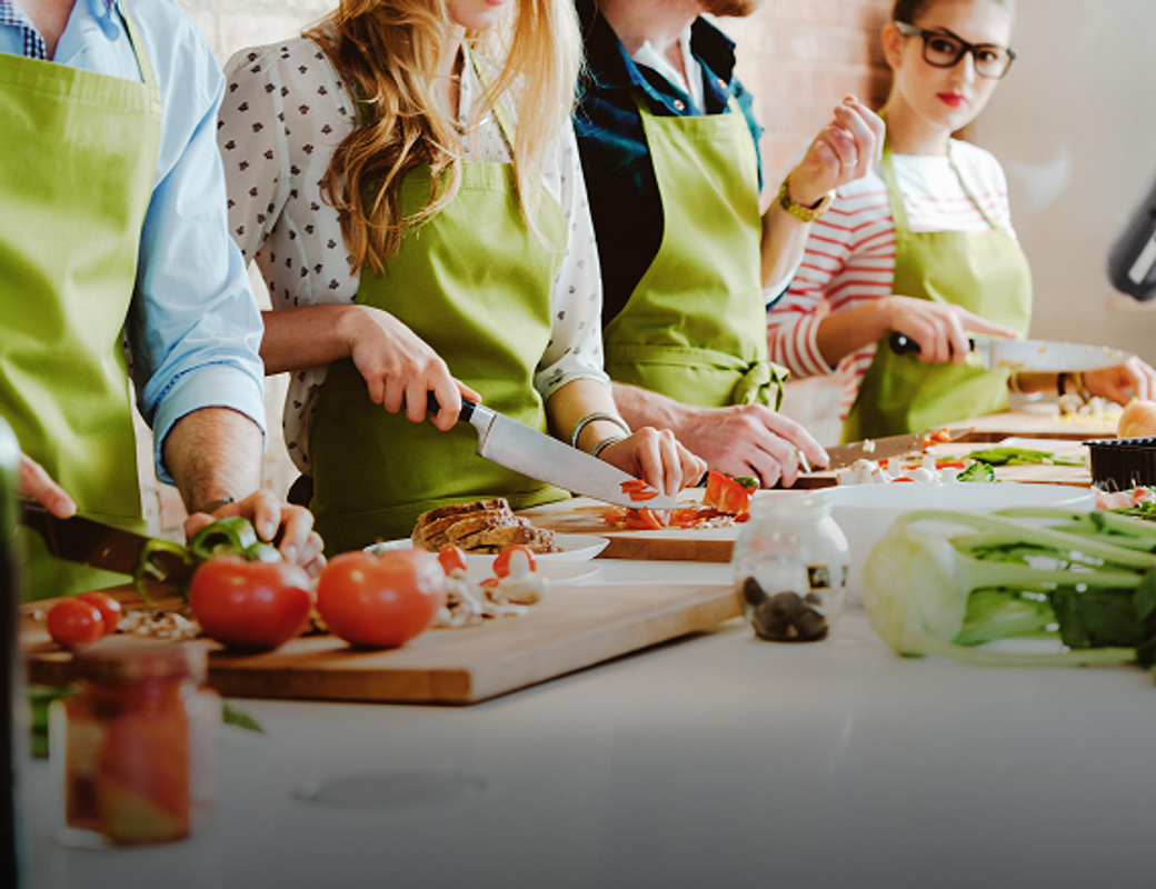 A group of four people, two men and two women, are in a kitchen wearing bright green aprons and chopping vegetables. The scene is brightly lit, and the focus is on the people and the fresh ingredients, suggesting a cooking class or a shared meal preparation.