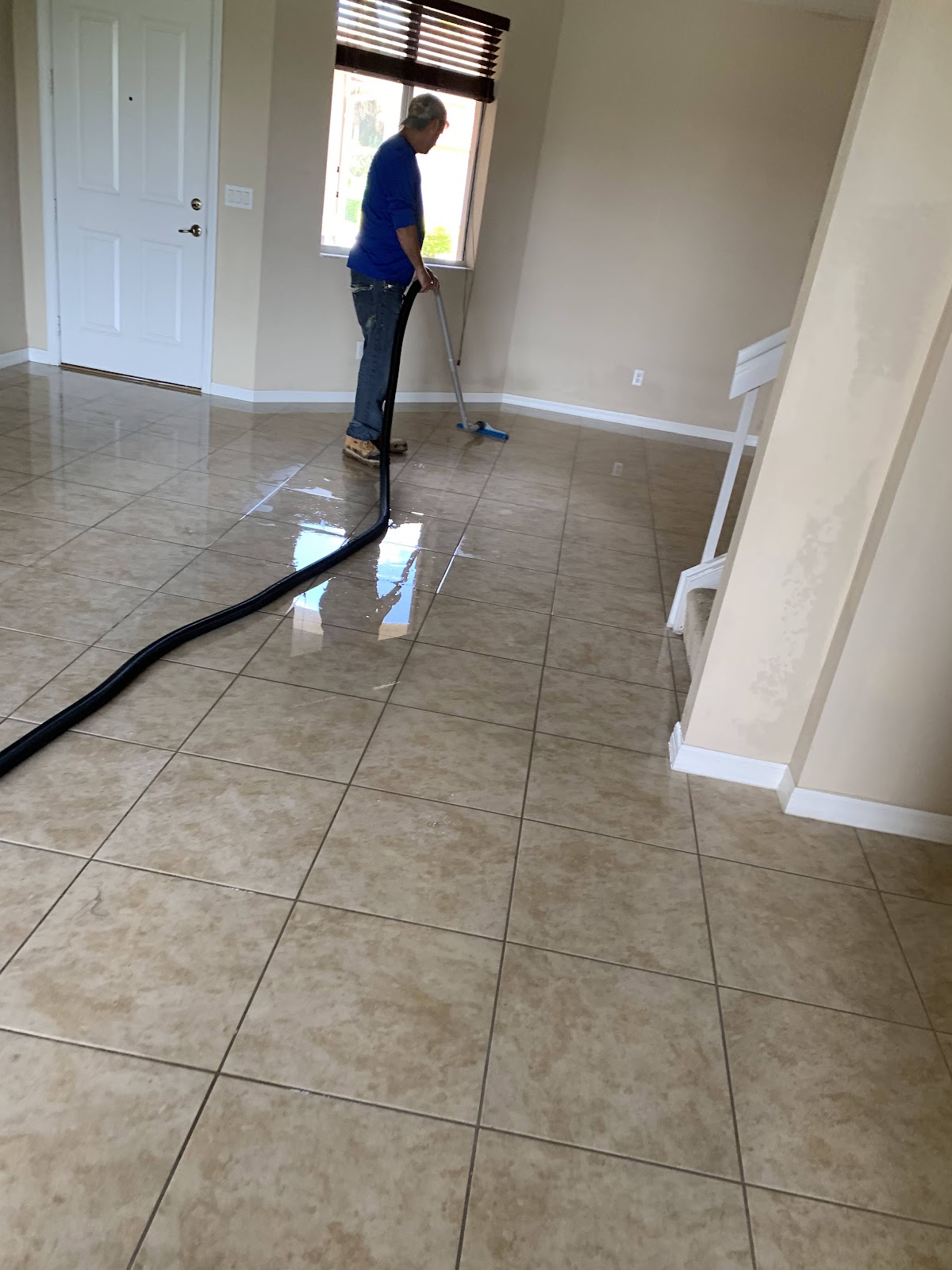 A man in a blue shirt and jeans is using a blue mop with a long handle to clean up a large amount of standing water on a tiled floor. A thick black hose runs across the floor from left to right. The room appears to be a living area or hallway, with a white door to the left and a window with blinds to the right. The walls are painted a light beige color.
