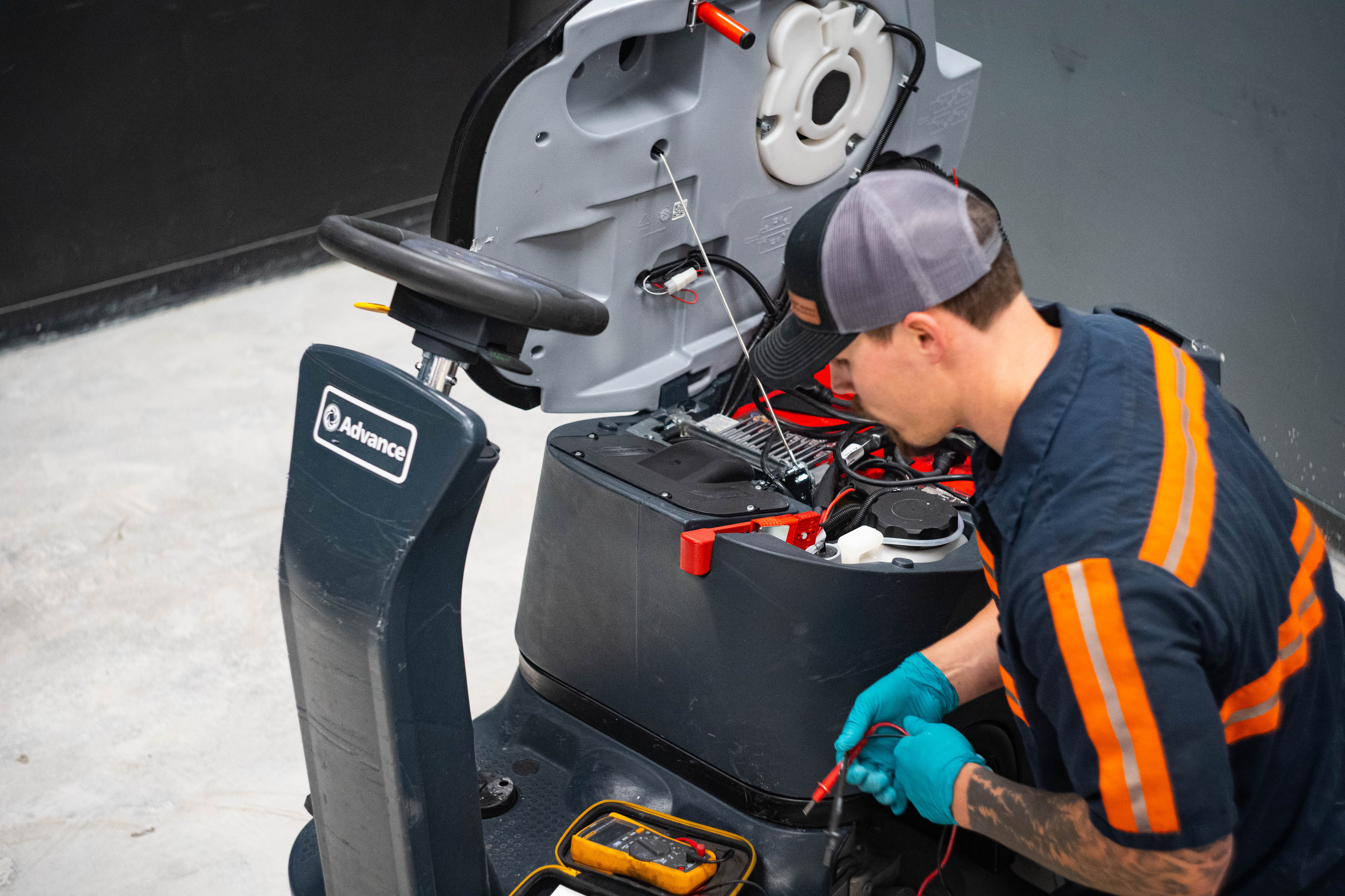 High-angle view of a WAY Solutions technician utilizing a digital multimeter to test the live circuitry and electrical components inside the battery bay of an Advance ride-on commercial floor scrubber.